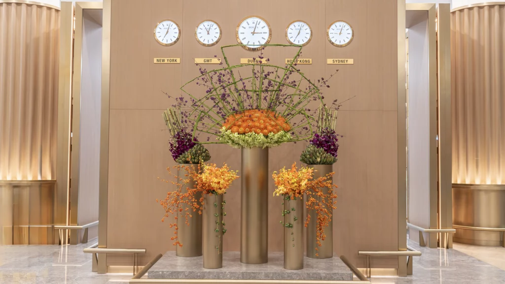 Large luxury floral installation with orange, green, and purple flowers arranged in tall gold vases under world clocks in a modern lobby.